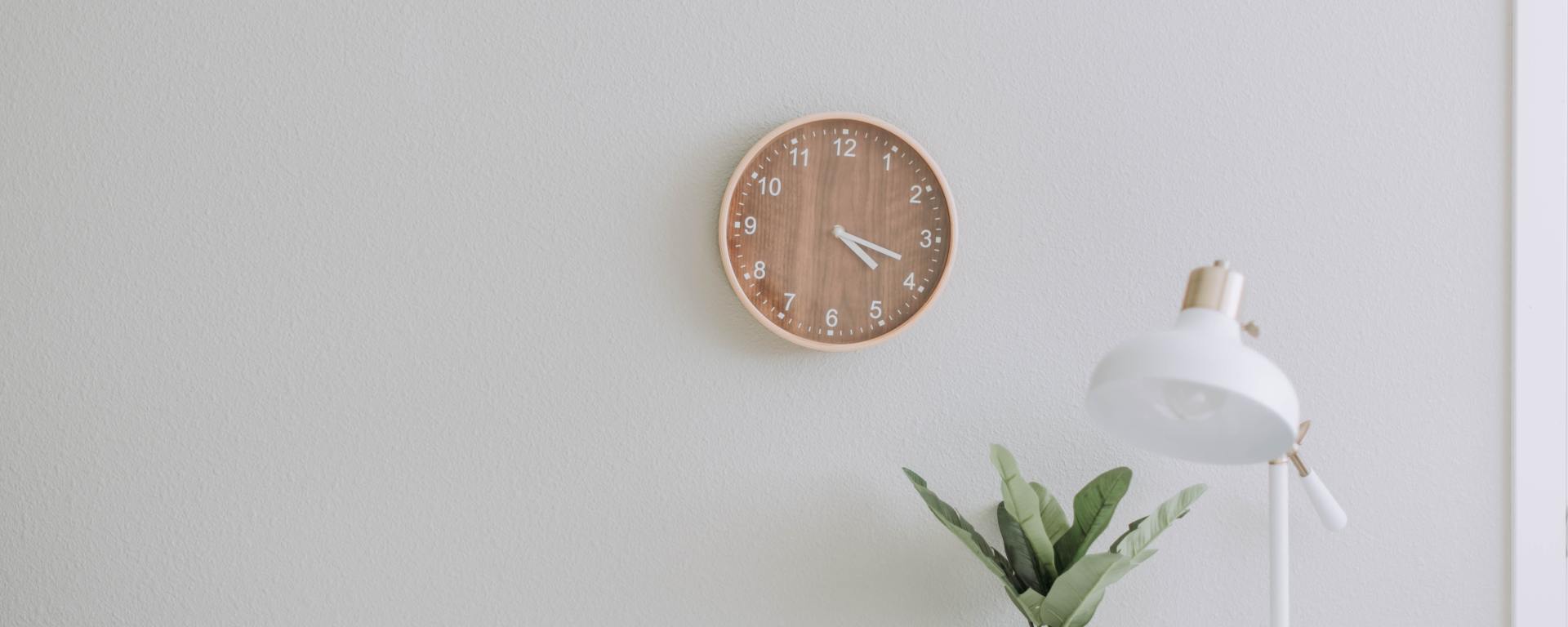 An image of a clock on a wall, a plant and lamp on a desk below.